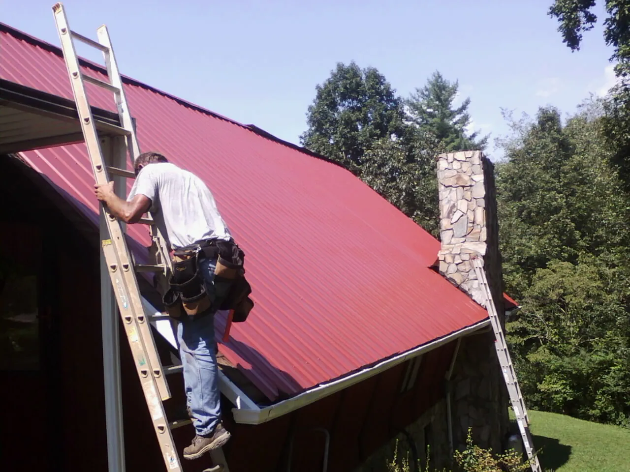 Roofer on a ladder beside a red metal roof and stone chimney — Soffit & Fascia Repair by Roofing Brothers in Pen Argyl